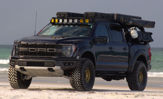 A Dark Blue Ford F-150 Raptor parked on a sandy beach with the ocean in the background. The truck is equipped with a roof-mounted light bar, spare tire, and cargo gear, and has large off-road tires with gold-colored wheel rims.