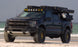 A Dark Blue Ford F-150 Raptor parked on a sandy beach with the ocean in the background. The truck is equipped with a roof-mounted light bar, spare tire, and cargo gear, and has large off-road tires with gold-colored wheel rims.
