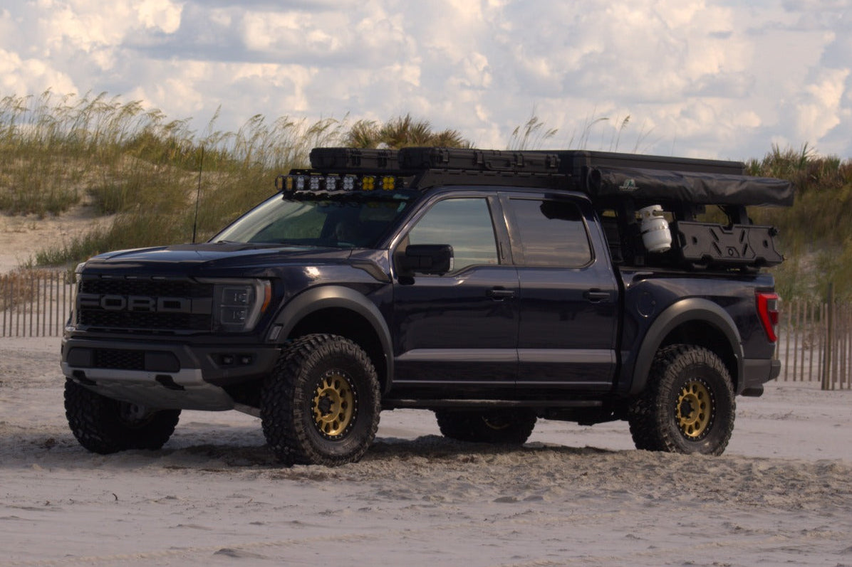 A Dark Blue Ford F-150 Raptor with a Aegon Ironwork roof-mounted camping setup, parked on a sandy beach with dunes and a cloudy sky in the background.