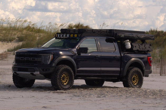 A Dark Blue Ford F-150 Raptor with a Aegon Ironwork roof-mounted camping setup, parked on a sandy beach with dunes and a cloudy sky in the background.