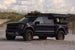 A Dark Blue Ford F-150 Raptor with a Aegon Ironwork roof-mounted camping setup, parked on a sandy beach with dunes and a cloudy sky in the background.