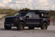 A Dark Blue Ford F-150 Raptor with a Aegon Ironwork roof-mounted camping setup, parked on a sandy beach with dunes and a cloudy sky in the background.