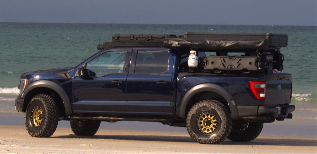 A dark blue off-road Ford F-150 Raptor with a Aegon Ironwork roof top tent and gear in the bed, parked on a sandy beach with the ocean and cloudy sky in the background.