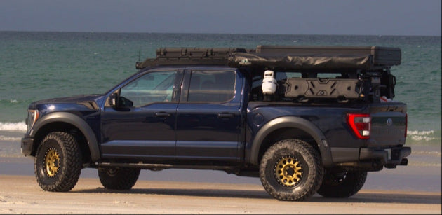 A dark blue off-road Ford F-150 Raptor with a Aegon Ironwork roof top tent and gear in the bed, parked on a sandy beach with the ocean and cloudy sky in the background.